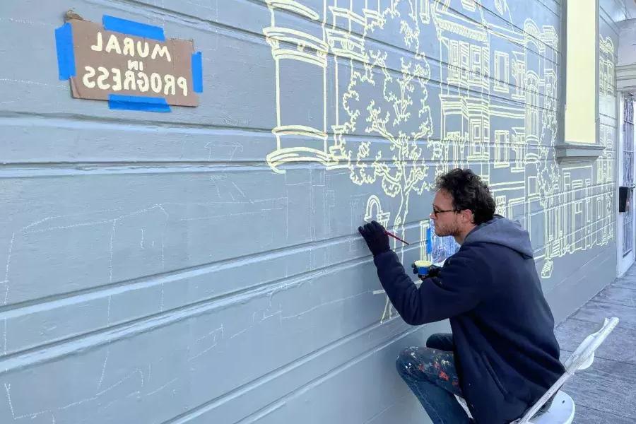 Ein Künstler malt ein Wandgemälde an der Seite eines Gebäudes im Mission District, an dem ein Schild mit der Aufschrift „Mural in Progress“ angebracht ist. San Francisco, Kalifornien.