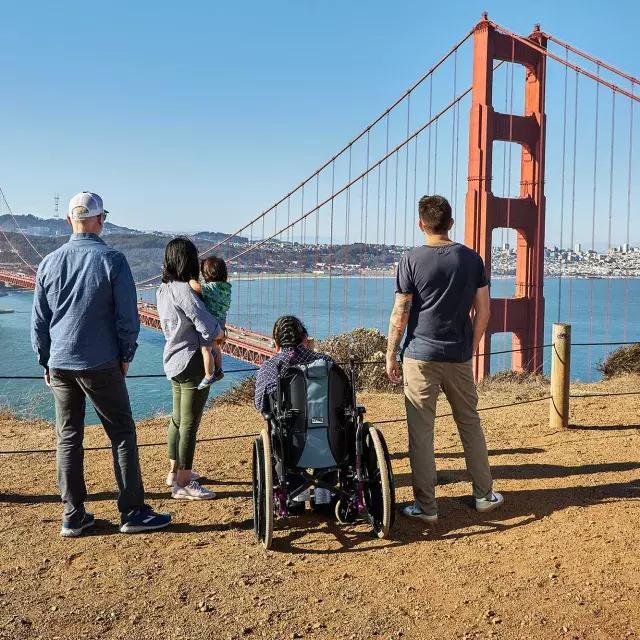 Un groupe de personnes, dont une personne en fauteuil roulant, est vu de dos alors qu'ils regardent le Golden Gate Bridge depuis Marin Headlands.