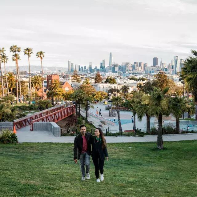 ドロレス公園と贝博体彩appのスカイラインを背にカメラに向かって歩くカップル。
